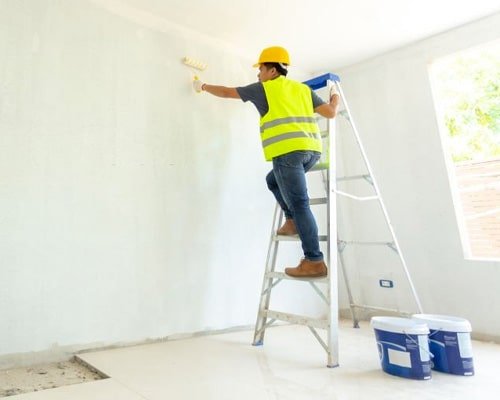 Pintor realizando su trabajo en una habitación subido en una escalera de tijera para alcanzar el techo